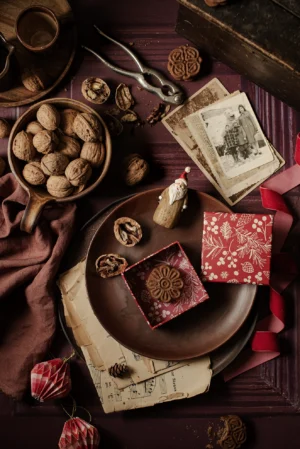 Tło do fotografii kulinarnej i produktowej Christmas is Coming. Kolor tła na którym jest stylizacja to ciemna czerwień, winny. Brązowy talerz z ciasteczkiem w czerwonym pudełeczku i figurka drewniana świętego Mikołaja. W brązowej misce są orzechy a obok leżą stare zdjęcia i dziadek do orzechów. Świąteczna scenka, którą domykają: czerwona wstążka i papierowa dekoracja.