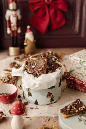 Tło fotograficzne Christmas is Coming które znajduje się w kadrze jako tło tylne. Czerwona welwetowa kokarda przyczepiona do tła wzmacnia świąteczny charakter zdjęcia. Stylizacja przestawia pudełko słodkości w klimacie świątecznym, Drobne kawałki pokruszone w koło pudełka a dodatkowe elementy w postaci foigurek nadają zdjęciu jeszcze bardziej świątecznewgo klimatu.