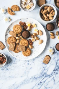 Tło fotograficzne Frosted Marble Wonderland na którym ułożony jest biały talerz wypełniony ciasteczkami i bakaliami. Stylizacja jest zimowa i dopelniają ją białe miseczki wypełnione orzechami i bakaliami.